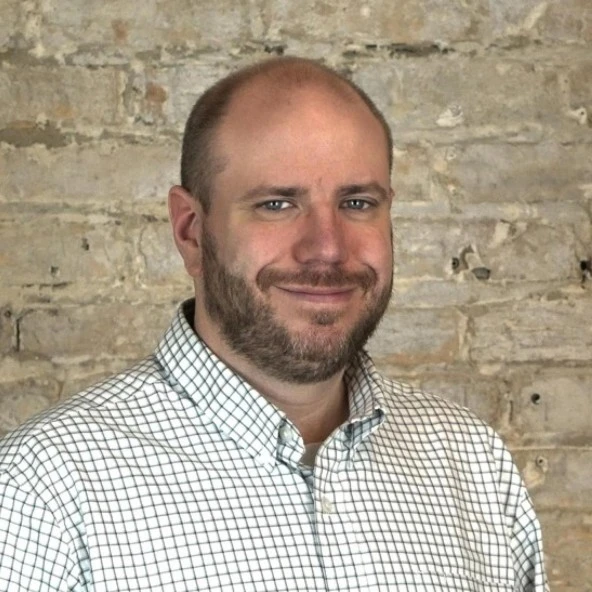Photo of James Voss Grumish appearing smart, but approachable, in a collared shirt but no tie; his brown hair is thinning and cut less than a quarter-inch on top; his beard is similarly shaded and short, but with noticeable strands of white; he stands in front of the interior exposed brick wall of a modern office in a renovated historic industrial building; the photo itself is clearly from a semi-professional photoshoot when his work colleague brought a nicer camera to the office.